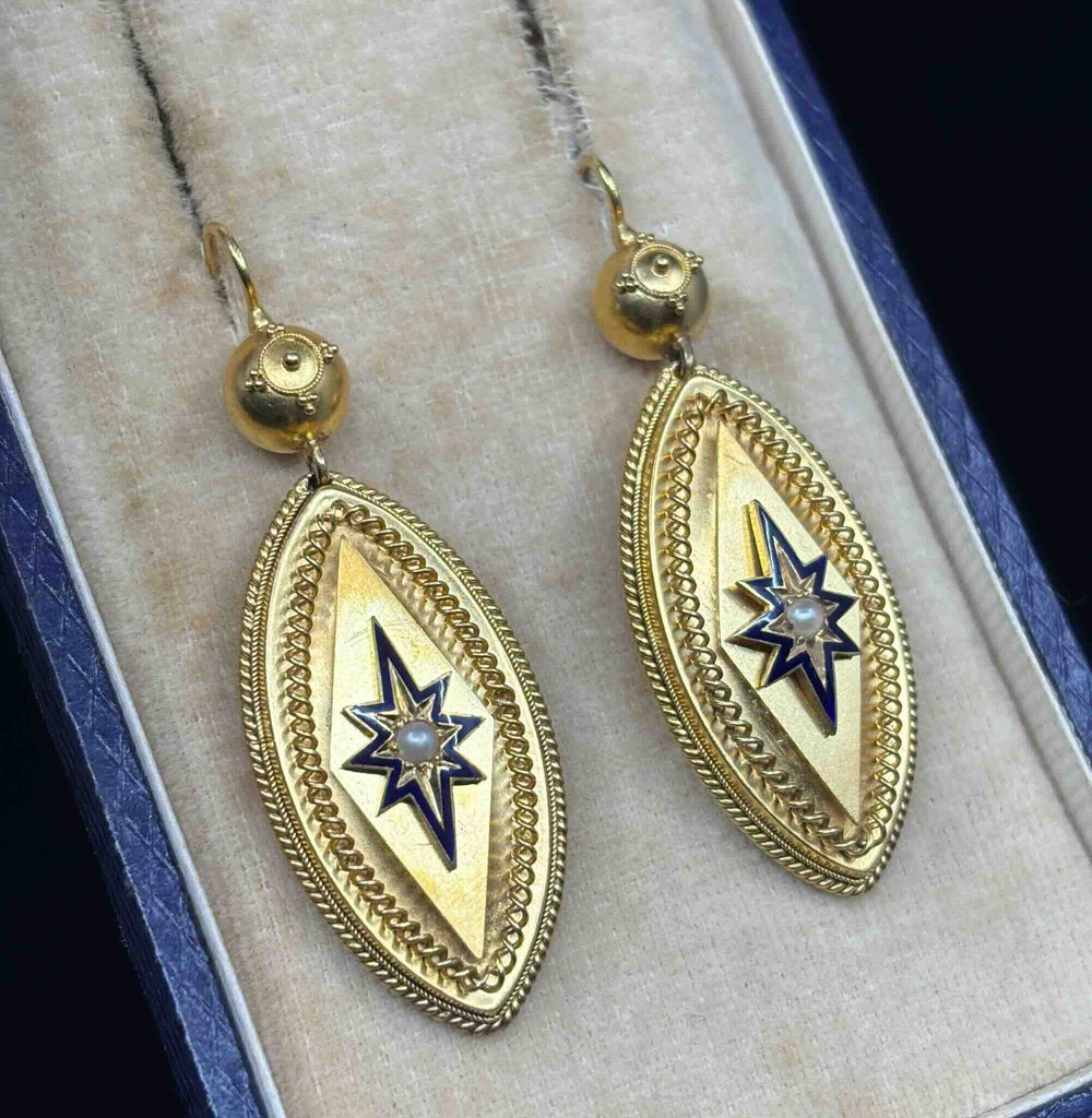 Angled close-up of antique Victorian gold earrings showing enamel starbursts, seed pearls and intricate cannetille borders