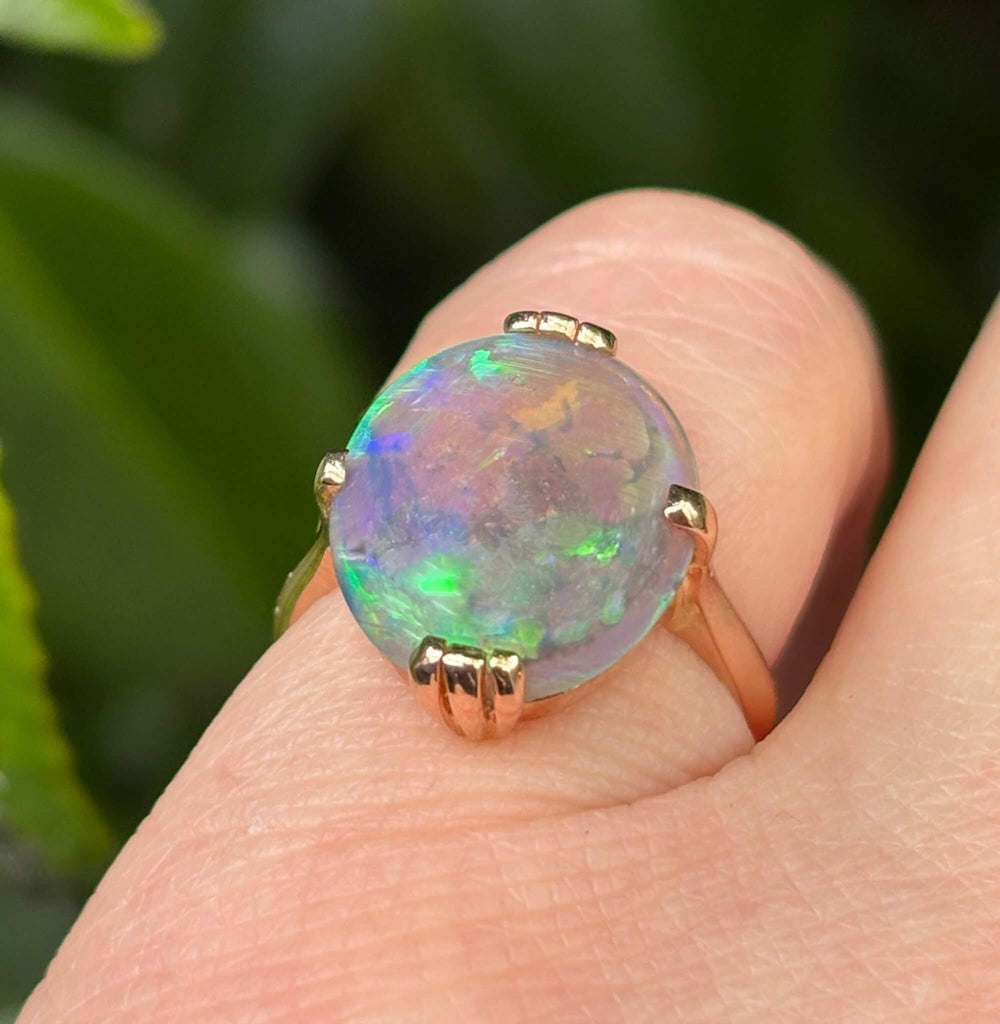A close-up of a gold ring with an oval, iridescent soild opal worn on a finger, set against a blurred natural green background.