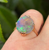 A close-up of a gold ring with an oval, iridescent soild opal worn on a finger, set against a blurred natural green background.