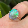 A close-up of a rose gold ring with an oval opal gemstone displaying vibrant green, blue, and purple flashes, worn on a finger against a natural green background.