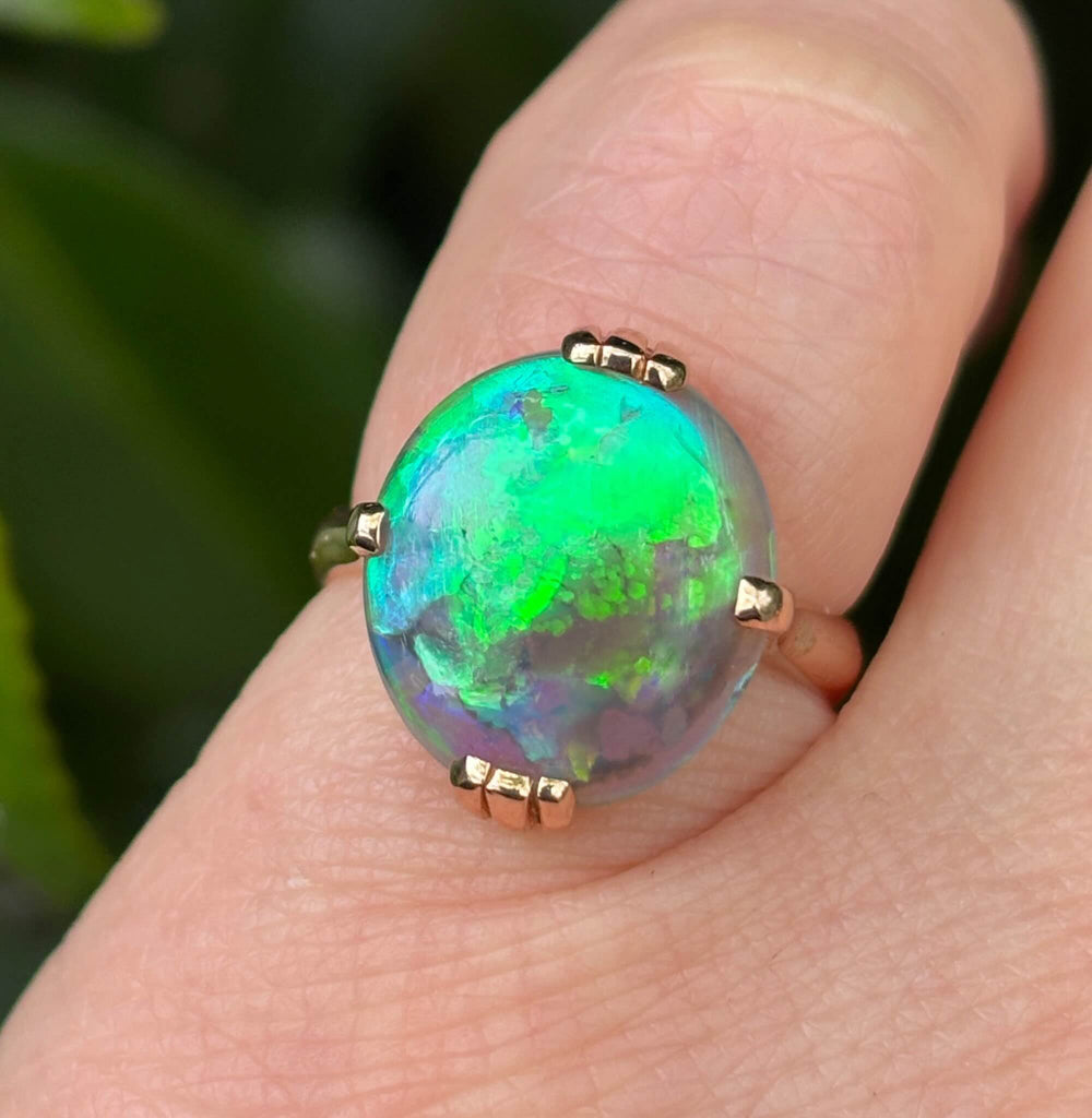 A close-up of a hand wearing a gold ring with a large oval cabochon opal that displays vibrant green, blue, and purple iridescence.