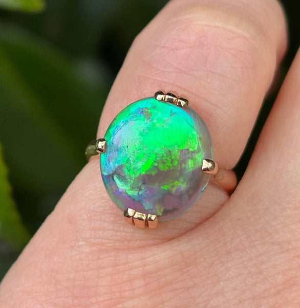 A close-up of a hand wearing a gold ring with a large oval cabochon opal that displays vibrant green, blue, and purple iridescence.