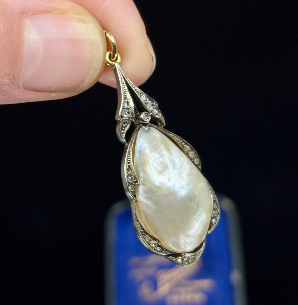 A close-up of a hand holding a pear shaped pearl pendant with rose cut diamonds set in silver and gold against a dark background with a blurred antique blue box below.