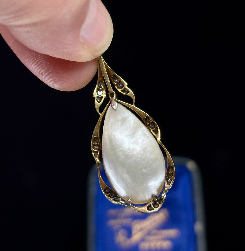 A close-up setting of the back of an antique gold pendant with a large pear shaped pearl with a blue box blurred in the background.