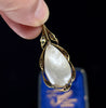 A close-up setting of the back of an antique gold pendant with a large pear shaped pearl with a blue box blurred in the background.