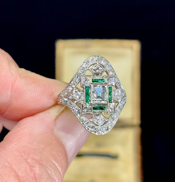 Stunning Art Deco diamond cluster ring featuring a rectangular cut diamond surrounded by emeralds and brilliant cut diamonds.