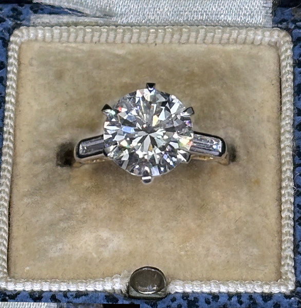 A round-cut diamond solitaire ring in a silver-toned band, displayed in an open beige velvet jewelry box.