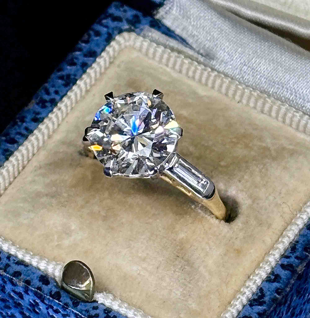 A round brilliant-cut diamond engagement ring with a white  and yellow gold band, displayed in a beige velvet ring box.