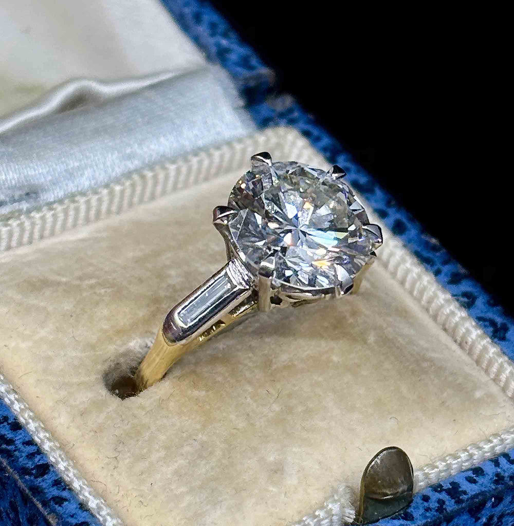 A round-cut diamond ring in a white and yellow gold solitaire setting, displayed in a cream colored velvet ring box with a blue velvet outer edge.