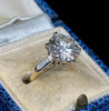 A round-cut diamond ring in a white and yellow gold solitaire setting, displayed in a cream colored velvet ring box with a blue velvet outer edge.