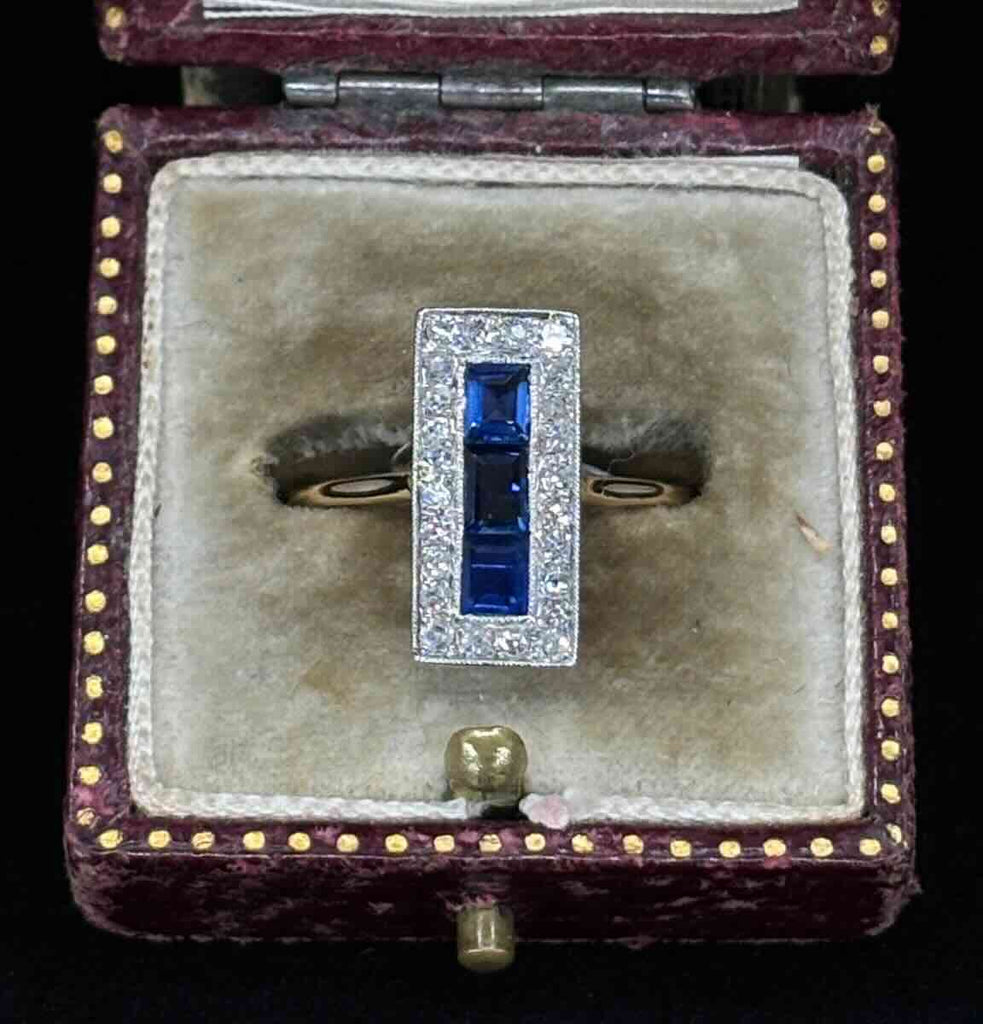 Front view of an antique Art Deco sapphire and diamond rectangular cluster ring held between fingers.