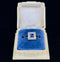 An elegant square sapphire engagement ring with a diamond halo, resting on a plush blue cushion inside a jewellery box.