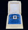 An elegant square sapphire engagement ring with a diamond halo, resting on a plush blue cushion inside a jewellery box.